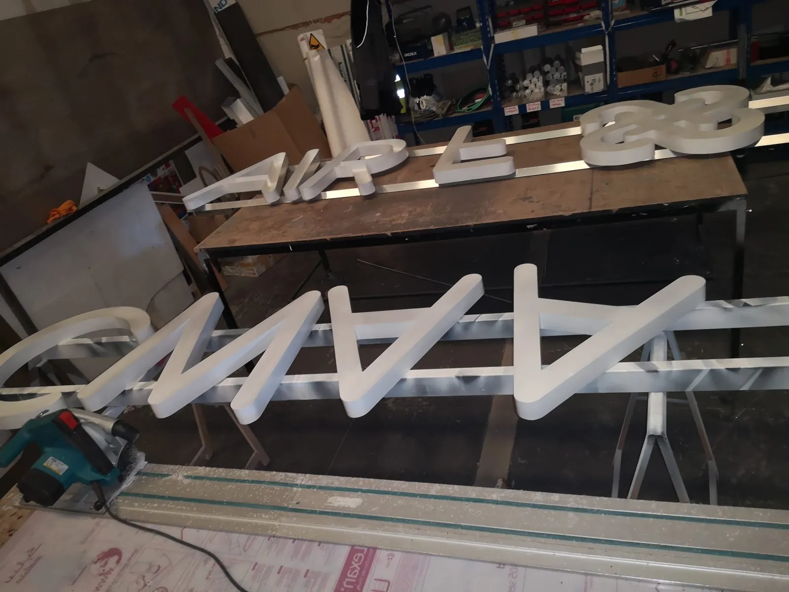 Letras corpóreas blancas de gran tamaño fijadas a perfiles de aluminio durante su fabricación, junto a una sierra de corte con carril guía